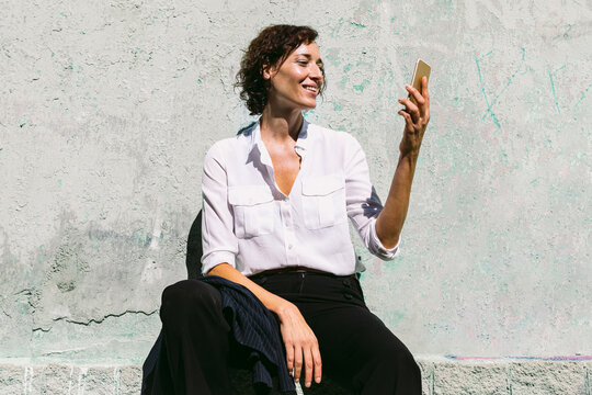 Smiling woman sitting on stone border and watching smartphone