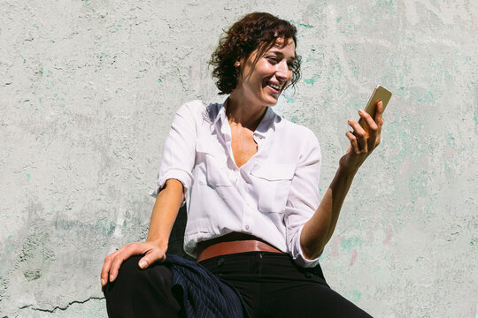 Smiling woman sitting on stone border and watching smartphone