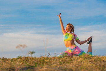 Young girl in bright tracksuit performs exercise to stretch muscles of legs while sitting on green lawn against background of blue summer sky. Hand is raised up.