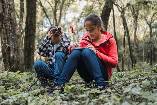 Ethnic siblings with binoculars and notebook exploring forest