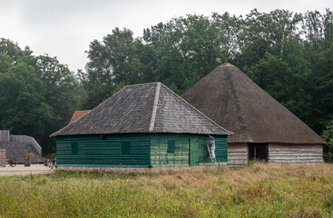 Fototapeta na wymiar Genk, Belgium - August 11, 2021: Domein Bokrijk. 2 barns, one is painted and has tile roof, while the other. the biggest has thick straw roof. Green meadow in front, darker green foliage in back.