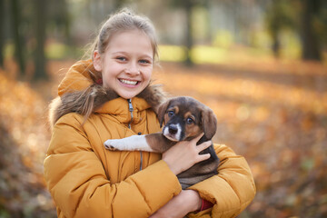 Front view of pretty girl in yellow jacket holding small dog on forest background. Concept of walking child in forest with dog.