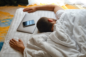 The man fell asleep in bed with a laptop and a telephone. Man working on laptop and phone in bed. Freelancer home office. Gadget addiction. Relax after work day concept