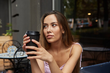 beautiful young asian woman with dark long hair, sitting at table outdoors with laptop, having rest. student having cup of coffee outside of cafe after doing homework. distant work, online education