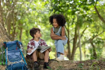 African American ethnicity boy and girl children wearing plaid shirt and backpack sitting at tree base talking and looking down on the map travel on hand in the park background 