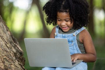 Naklejka na ściany i meble children girl African American ethnicity black skin sitting on tree base use Laptop computer to chat with friends via video call with 5G internet signal in the park