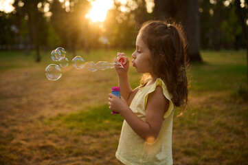 Adorable child baby girl, blowing soap bubbles at sunset, enjoying pleasant time outdoors in the meadow. Sun's rays fall through transparent bubble spheres with iridescent reflections.