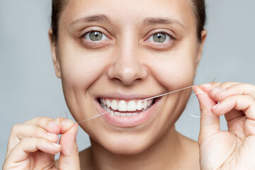 A close-up of a young beautiful caucasian woman flossing her teeth isolated on a gray background....