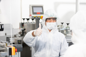 male engineers wearing personal protective equipment uniform(PPE) wearing medical face mask, producing medical face masks thumbs up pose beside machine in laboratory