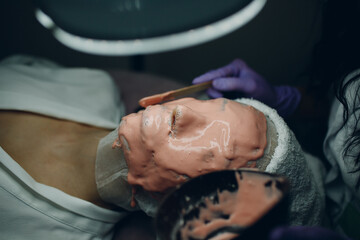 Obraz na płótnie Canvas Alginate face mask application process. A female hands of a beautician holding spatula. Woman lying on a couch in office of cosmetologist. Facial skincare in the beauty spa salon.