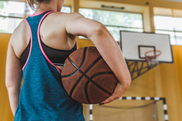 Female high school basketball player holding a ball on school gymnasium, with basketball board in background.