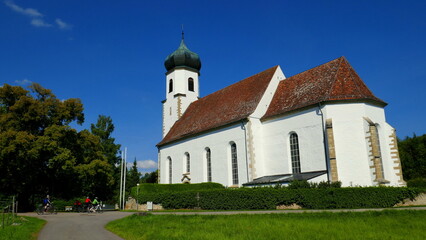 Fototapeta premium sehr schöne Dorfkirche in Poltringen auf grüner Wiese mit Bäumen unter blauem Himmel und Radfahrern