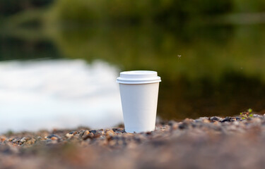 A white paper cup of coffee or tea stands on the rocky shore of the lake. A mug of hot drink on a pebble. Beautiful nature background of the lake in the forest behind. free copy space.