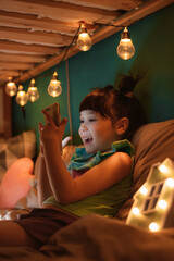Little happy girl with funny hairstyle sitting in childrens room on her bunk bed holding smartphone