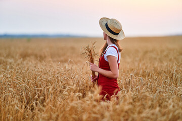 Calm serene free young female wearing straw hat and red denim overall standing in golden yellow dry wheat field and enjoying beautiful freedom moment