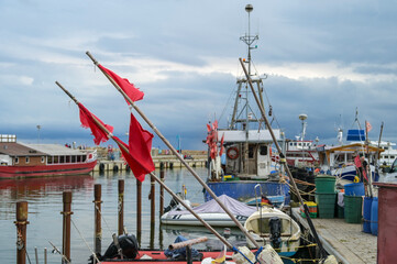 Fototapeta na wymiar Red flags and fishing boats at the dock in the port of Sassnitz on the island of Rugen in the Baltic Sea against a cloudy sky, travel and tourist destination, copy space, selected focus