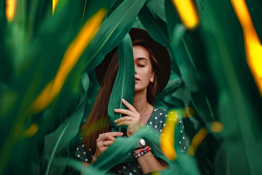Portrait of a sensual stylish fashionable young beautiful woman with smooth face skin, closed eyes and natural makeup wearing hat and polka dot dress holding green leaf in her hands