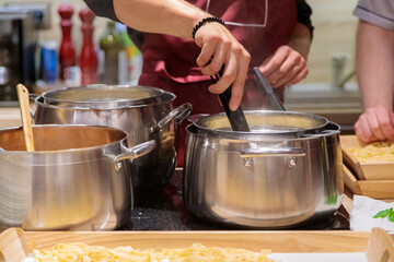 The chef prepares pasta in large metal pans. Male hands take out the pasta from the pan with tongs. Close-up.