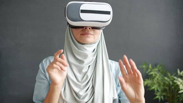 Slow motion of cheerful Middle Eastern woman in hijab using virtual reality glasses moving arms enjoying cyber space indoors at home. Technology and fun concept.