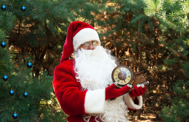 Santa Claus in a red suit against the background of green Trees of the forest in his hands holds an old wooden clock that shows 6 hours with space for text at Christmas