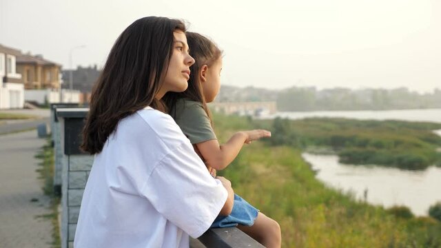 Careful mother holds hand of Asian little girl sitting on large parapet pedestal on contemporary city embankment at sunset, side view