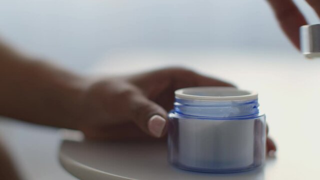 Woman opening blue glass jar of nourishing cream to protect skin, taking cream and applying in on hands, close up