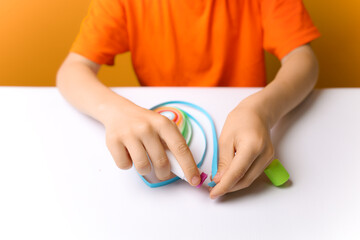 a boy in an orange T-shirt sits at a white table and prepares thin strips of paper for gluing. the child learns the technique of quilling