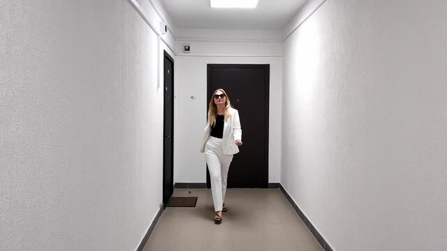 White woman in white suit confidently walks down a white corridor toward camera