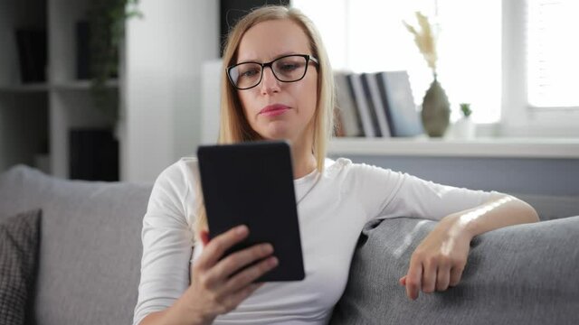 Beautiful woman in eyeglasses and casual wear having video conversation on digital tablet. Caucasian blonde sitting on cozy sofa and using modern device.