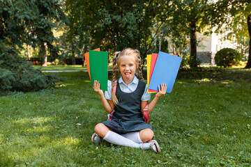 Education concept. Cute smiling schoolgirl sitting on the grass. Happy little girl holding books - obrazy, fototapety, plakaty