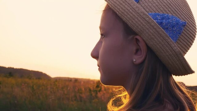 A beautiful teenager girl in a straw hat with a blue ribbon, in the rays of the setting sun, stands on a meadow field. Looks into the distance thinking about a wonderful future.