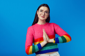Attractive cheerful girl keeps palms pressed together in praying gesture, wears rainbow sweater,...