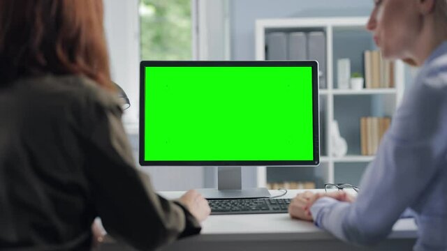Two business women in formal clothes sitting together at desk and using modern computer with green empty screen. Concept of people, technology and cooperation.