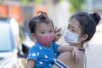 Senior Asian woman wearing surgical face mask is holding her nephew (Grandson) with red cloth mask and help to adjusting his mask to prevent coronavirus while walking outside.