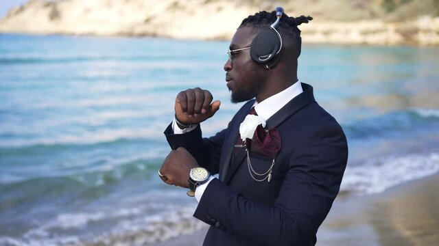 Carefree handsome African American elegant man in headphones dancing in slow motion on Mediterranean sea coast. Portrait of happy relaxed male tourist on sandy beach with foamy waves rolling