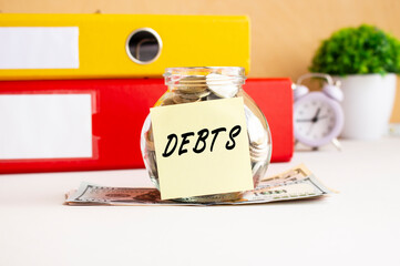 A glass jar with coins stands on a stack of dollar bills on the desktop. The jar has a sticker with the text DEBTS.