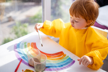 Toddler paints a rainbow on paper sitting at home.Flash mob society community on self-isolation...