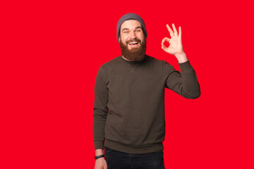 Naklejka premium Bearded hipster wearing warm clothes is showing ok gesture over red background.