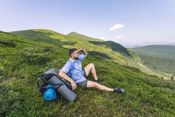 Naklejka na ściany i meble mountain hiking concept. a guy with a big backpack squatting to rest. hiker on top of a mountain