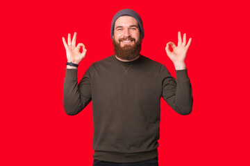 Pleased bearded man is showing ok gesture over red background while smiling at the camera.