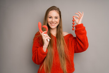 An attractive girl in a sweater depicts the emotion of happiness, smiling.holding a red dildo, vaginal balls in her hand - obrazy, fototapety, plakaty