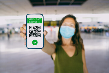Young girl in the airport wearing a face mask, holding a smartphone with a Green Pass. The Green Pass give access and safety in the airport. Health Passport concept. Focus on the smart phone.