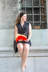 Dressing in black sleeveless clothes a pretty woman is reading a red book outside by a window..