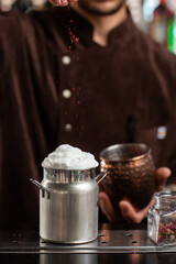 Barman gently decorating cocktail in metallic glass. Body of bartender in brown apron on background.