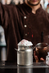 Barman gently decorating cocktail in metallic glass. Body of bartender in brown apron on background.