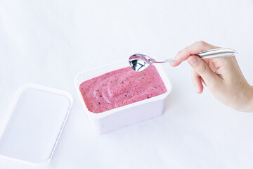 Berry ice cream pink color cream texture in a rectangular white container with an open lid and hand with metal spoon on a white background. Top view. Eating concept