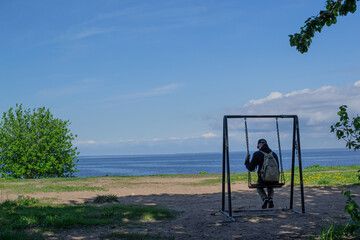 Sad adult guy, a man in a cap and with a backpack sits on a swing and sways among the green trees against the background of the blue sea. Back view