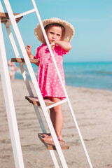 A little girl in a pink dress and a straw hat on her head stands on the stairs on the beach against the background of the sea, rescue point
