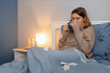 Cute girl in beige sweater sitting in bed under blanket and drink remedy for heat temperature. Woman feeling sick. Symptoms of cold and flu. Seasonal disease. Near paper napkins for blowing nose.