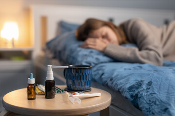 Obraz na płótnie Canvas Selective focus on various pills, spray for a runny nose, thermometer and cup with hot beverage. On the background young girl sleep in bed. Remedy for disease. Woman feeling sick. Treatment of cold.